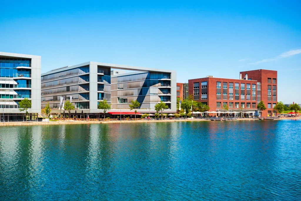 Contemporary buildings along a riverfront with trees and outdoor seating under a bright blue sky.